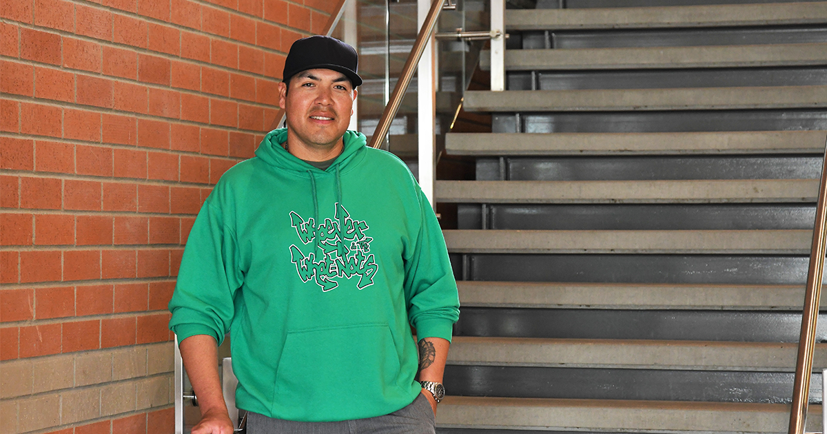 A man is standing in front of a staircase looking at the camera. He is wearing a green hooded sweatshirt and a black hat. He has both hands in his pockets.